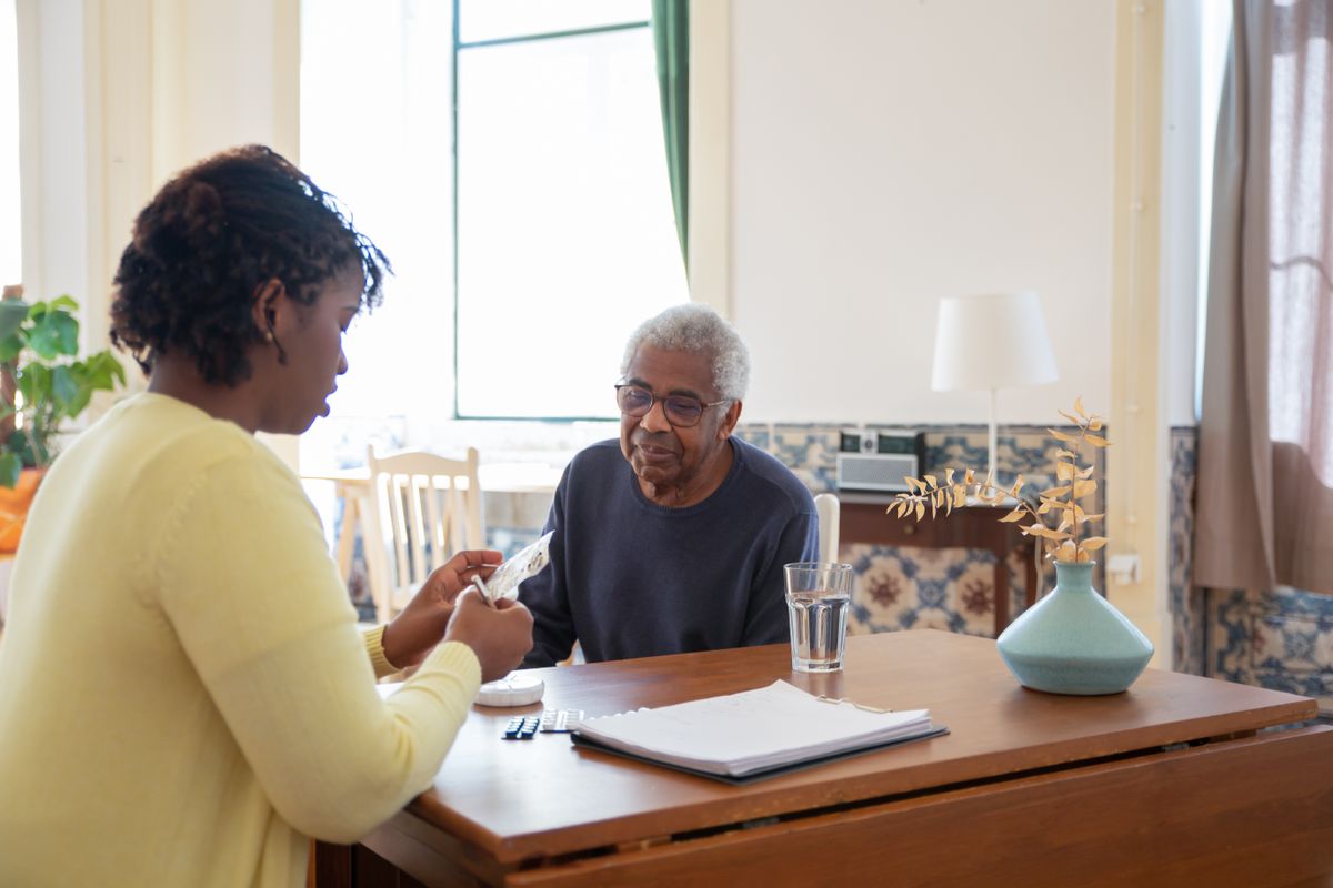 A Nigerian caregiver helping an older man with his medication at a sunlit dining table